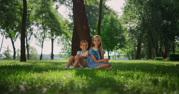 Smiling brother sister play on green grass under tall tree. Happy children sitting on lawn summer day. Little blondhair siblings resting on nature sunny weekend. Kids have fun together outdoors.