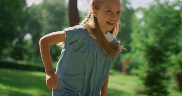 Smiling beautiful girl running on soft green grass outdoors. Happy little lady playing active game on summer nature. Cute blondhair child have fun in sunny forest. Playful kid jogging on sunlight.