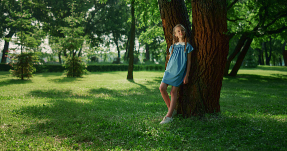 Dreamy girl lean tree trunk in park. Smiling teen observing meadow nature. Cute child in blue dress imagining fantastic stories. Girl enjoy warm summer day alone. Carefree childhood concept.