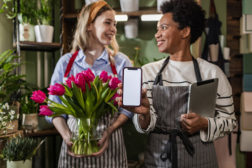 Portrait of two multiracial flower store workers looking at each other. A young blond woman is holding a flower bouquet while a Latin American woman is holding a digital tablet and showing a smart phone with a blank screen to the camera. Chroma key. Space for copy.