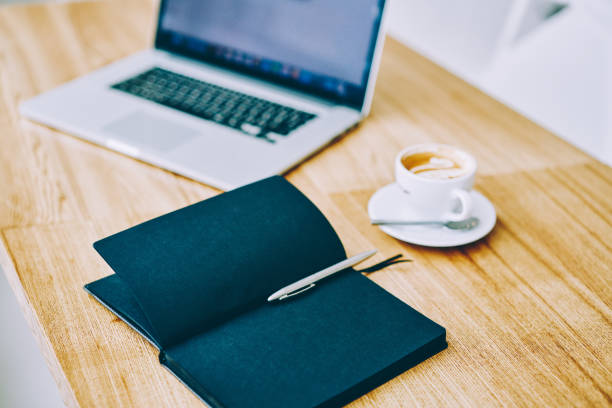 Opened notebook with black pages and pen placed on wooden table with laptop and cup of coffee saucer and spoon