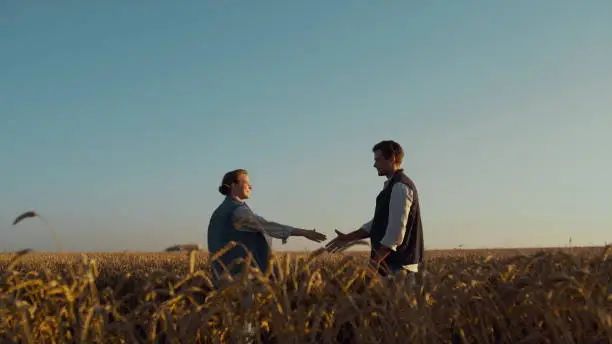 Farmers meeting wheat field in golden sunlight. Agronomists discussing agreement. Two agricultural business partners shaking hands sign papers on harvesting season deal. Successful cooperation concept