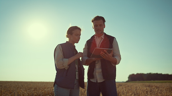 Agronomists team holding tablet computer in sunlight. Agritech industry workers. Smiling man farmer using pad discussing harvesting plan with farmland owner. Professional engineers work at wheat field