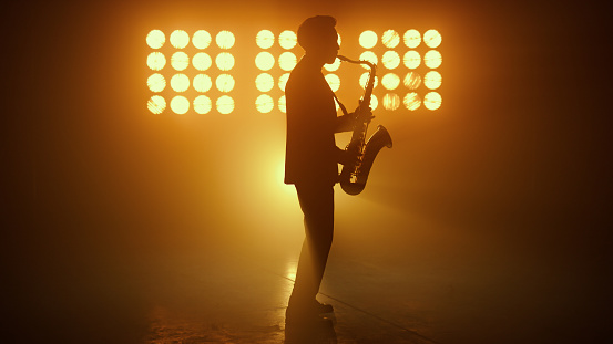 Silhouette of jazz musician playing saxophone in night club spotlights. Side view of unrecognizable stylish man performing melody on sax lit stage lights. Young guy saxophonist enjoying music indoors.