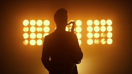 Unknown sax player making jazz show on nightclub stage. Silhouette of man musician playing saxophone on music concert indoors. Expressive young saxophonist performing melody on musical instrument.