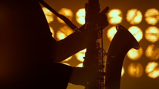 Unknown saxophonist hands playing musical instrument solo in studio spotlights close up. Professional saxophone player performing on stage night club. Silhouette of man musician holding sax indoors.