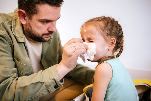 Dad wipes his sick child's nose with a white disposable paper napkin at home.