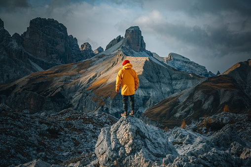 Rear view of a young hiker man in a yellow raincoat and a big backpack and outdoor sports eyewear standing on top of a rock hiking and walking in a fantastic mountain landscape on another side of Dolomiten mountains and a blue pond view of Drei Zinnen or Tre Cime di Lavaredo with a beautiful cloudy sky in South Tirol, Dolomites, Italia. Popular travel and hiking Destination in the world.