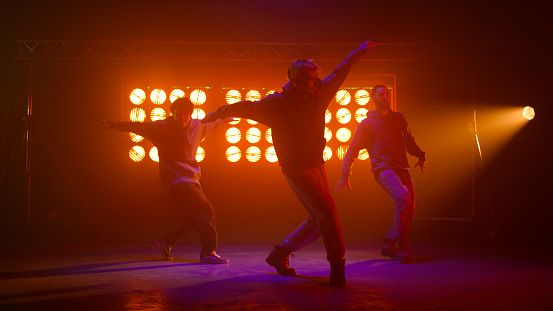 Hiphop performers moving body rhythmical on night club stage. Stylish guys performing emotionally wearing sport clothes on dancefloor. B-boys band dancing freestyle in flashing bright spotlights.
