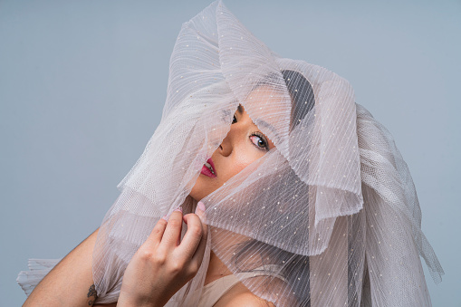 Average age 25-year-old Latina woman dressed in a beige and tulle bodysuit that she pulls over her head and covers her face in the same color stands inside a photo studio while looking towards the camera