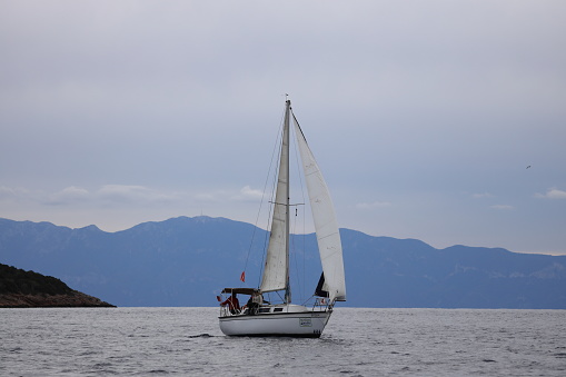 Bodrum,Mugla, Turkey. 04 February 2023: sailor team driving sail boat in motion, sailboat wheeling with water splashes, mountains and seascape on background. Sailboats sail in windy weather in the blue waters of the Aegean Sea, on the shores of the famous holiday destination Bodrum.
