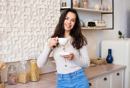 Portrait of happy woman posing in kitchen interior with cup of herbal tea and smiling at camera, enjoying morning coffee at home, free space