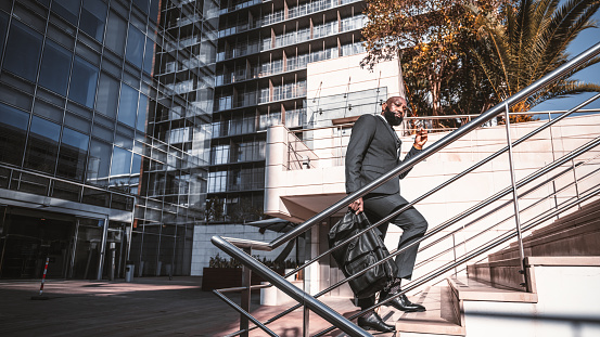 An African white-collar worker left the office's contemporary windowed building and is about to climb the stairway with a metal railing while carrying a weekender bag, with eyeglasses in his hand