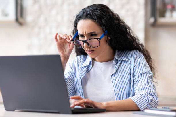 problemi di vista. signora latina che usa gli occhiali mentre lavora sul computer portatile a casa, seduta in cucina - strizzare gli occhi foto e immagini stock