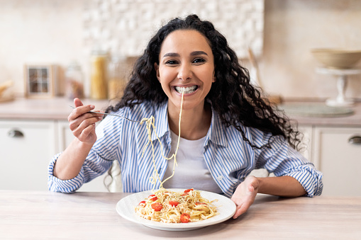 Happy excited latin woman eating tasty homemade pasta and smiling at camera, sitting at table in kitchen interior. Recipe of delicious spaghetti