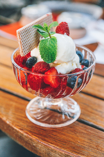 Tasty gelato summer dessert with strawberries, blueberries vanilla and chocolate sauces in glass decorated by mint and wafer on wooden restaurant table background
