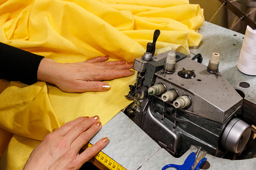 A close view of an experienced female dressmaker is using yellow textiles and a sewing machine for making new clothes.