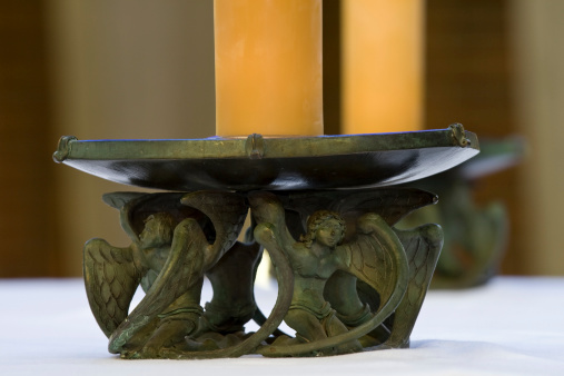 A matching pair of large-diameter, golden wax candles sit atop large candle holders adorned with angelic figures on a clean, white cotton tablecloth within an abbey run by benedictine monks.