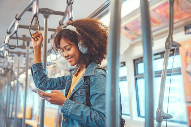 기차에서 스마트 휴대폰을 통해 음악을 듣는 행복한 젊은 아프리카계 미국인 여성 승객, 트램, 라이프스타일, 교통에서 이동하는 동안 무선 헤드폰을 착용한 미소 여성. - bus commuter passenger mobile phone 뉴스 사진 이미지