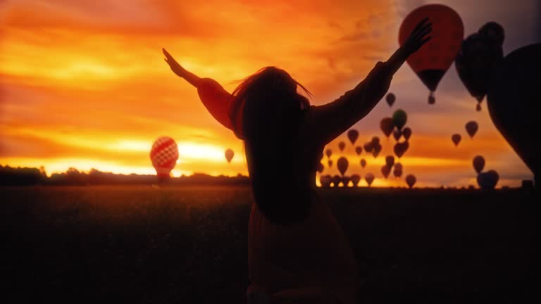 SLO MO Silhouette of a carefree woman in the field with hot air balloons in the sky at golden hour