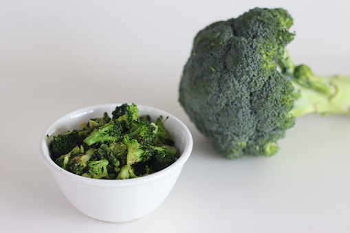 Sauteed broccoli in a white bowl, shot on a white background along with fresh bunch of broccoli