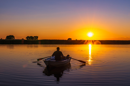Silhouette of a 60-year-old Caucasian man with a fishing rod and a poodle dog in a wooden rowing boat with oars in his hands against the background of a sunset.