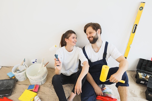 Happy young caucasian couple holding paint rollers during renovation on living room while sitting on floor in their new house
