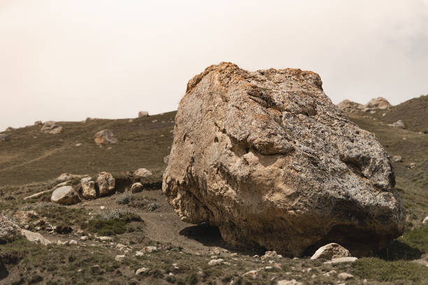 Mountain landscape - huge grey boulder with orange lichen, cracks, splits on slope with white sky, poor green grass in sunlight in highland in overcast summer weather. Travel in Caucasian mountain.
