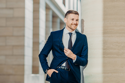 Image of successful businessman in elegant clothing, keeps hand in pocket, holds sunglasses, smiles cheerfully, looks somewhere, being confident in his business deal. People and job concept.