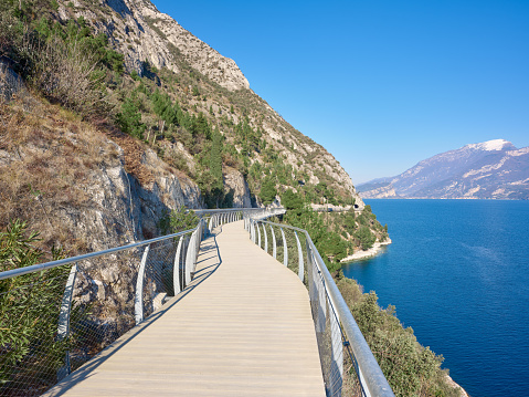 Wide-angle view of the hanging bicycle and pedestrian lane which runs from the town of Limone sul Garda to the border of Trentino region (municipality of Riva del Garda). The cliffs and slopes of the mountain range of Monte Baldo can be seen on the background. A perfectly clear sky, the warm light of a late afternoon, steep cliffs rising from the blue lake water and dotted with trees. High level of detail, natural rendition, realistic feel. Developed from RAW.