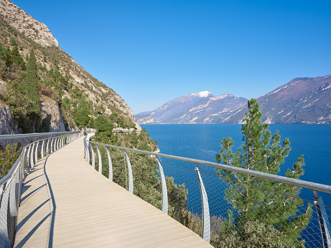 Wide-angle view of the hanging bicycle and pedestrian lane which runs from the town of Limone sul Garda to the border of Trentino region (municipality of Riva del Garda). The cliffs and slopes of the mountain range of Monte Baldo can be seen on the background. A perfectly clear sky, the warm light of a late afternoon, steep cliffs rising from the blue lake water and dotted with trees. High level of detail, natural rendition, realistic feel. Developed from RAW.