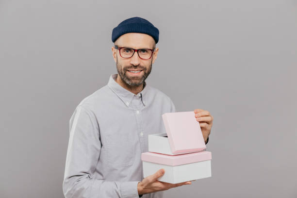 istock Handsome European man with thick stubble, pleasant smile, holds boxes in hands, opens gift with happiness, wears formal white shirt and headgear, isolated over grey background. Parcel in mans hands 1467244649
