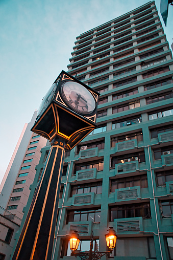 A vertical high angle shot of a metal street clock and a green tall building in the background in Macau, China