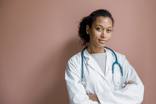 Portrait of female healthcare worker standing against brown wall. Young woman doctor in lab coat with stethoscope looking at camera with her arms crossed.