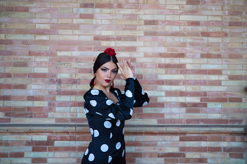 A Hispanic brunette flamenco dancer wearing a white polka dots dress against brown brick wall in Seville