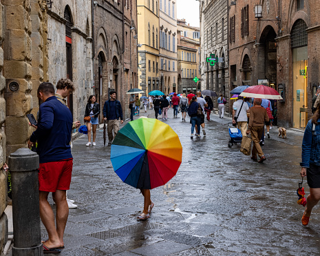 Sienna, Italy – July 25, 2021: A girl with a rainbow umbrella walking on a cobbled street in Sienna on a rainy day