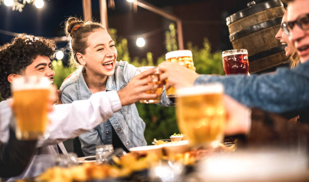 Happy friends clinking and toasting beer at brewery restaurant patio - Life style and beverage concept with young people having fun together out side at patio garden by night - Warm evening filter stock photo