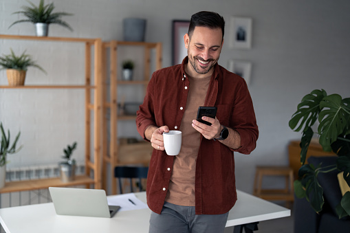 Cheerful businessman in casual clothes standing in front of table with laptop and paper document on it in home office, smiling, looking at smartphone and holding cup of coffee. Potted plants on shelves in background.