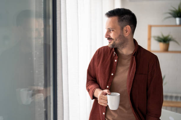 istock Satisfied smiling man holding white cup of coffee and looking away through window 1467275426