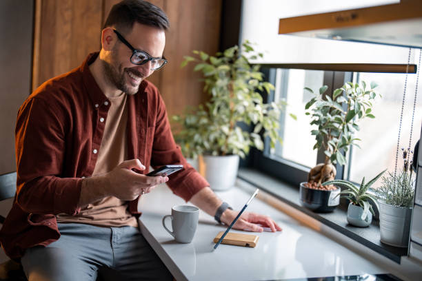istock Smiling man using smartphone sitting near window with potted plants 1467275436