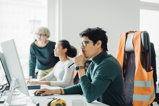 Young male architect sitting at his desk and working on computer with female colleagues working in background at office. Male and female professionals working in an engineering office.