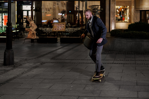 A young man with a briefcase is enjoying himself in the city at night while riding on a skateboard in a street.