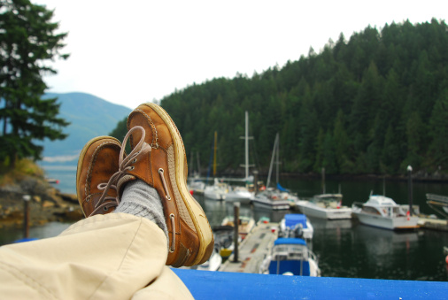 A pair of legs and deck shoes with marina and boats in background, selective focus on shoes