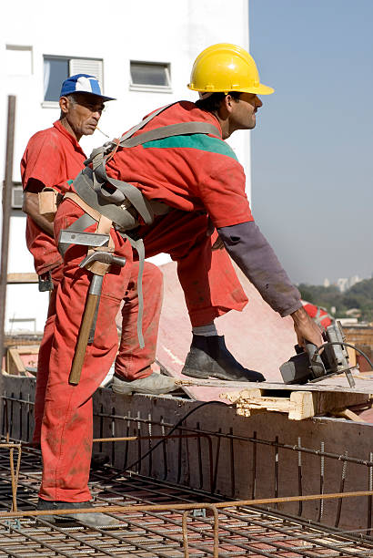 Two construction workers are working on the rooftop of an unfinished house.  The are both carrying tools, and one is wearing a hard hat.  They are both looking away from the camera.  Vertically framed shot.