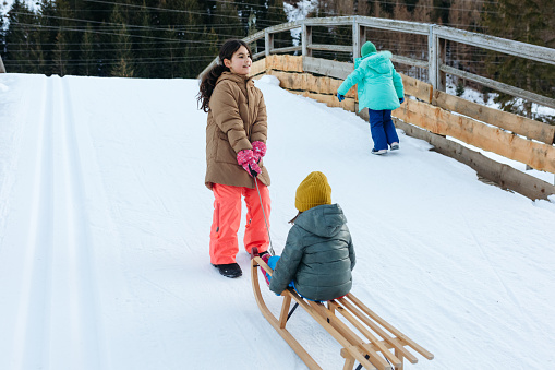 three kids going up to snow slope with small sister on wooden sled