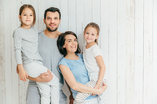 Portrait of happy parents carry their beautiful daughters on hands. Lovely family of four pose together against white background have positive expressions. Affectionate mother and father with children