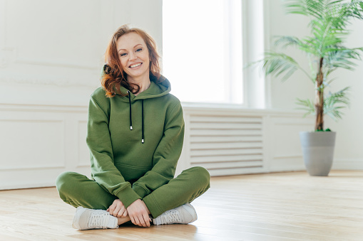 Horizontal shot of happy redhead sportswoman in green tracksuit, white sneakers, sits in lotus pose, has friendly smile on face, poses indoor. Female pilates instructor practices stretching exercises