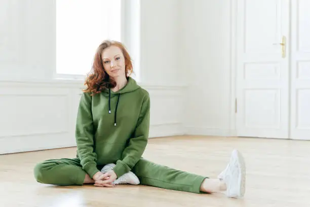 Indoor shot of glad ginger European female wears sports clothes, stretches legs on floor, wears sportshoes, has slim body, has appealing appearance. People, motivation and gymnastics concept.