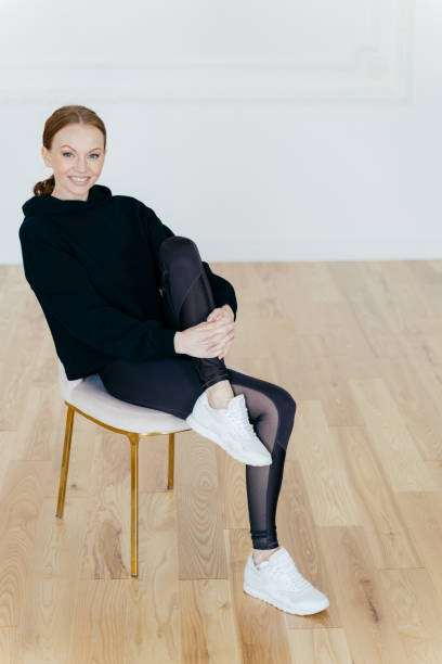 Satisfied woman with ginger combed hair, has pleasant smile, raises leg and keeps hand on knee, poses on chair in studio, has rest after aerobics, wears black sport costume and sneakers. Motivation mujer satisfecha con cabello peinado de jengibre, tiene una sonrisa agradable, levanta la pierna y mantiene la mano en la rodilla, posa en la silla en el estudio, descansa después de los aeróbicos, usa traje deportivo negro y zapatillas de deporte. motiv
