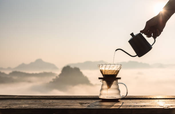 istock Hand pouring hot water on coffee ground with fillter, barista using drip coffee kit set with mountain and fog in sunrise background. 1467327599